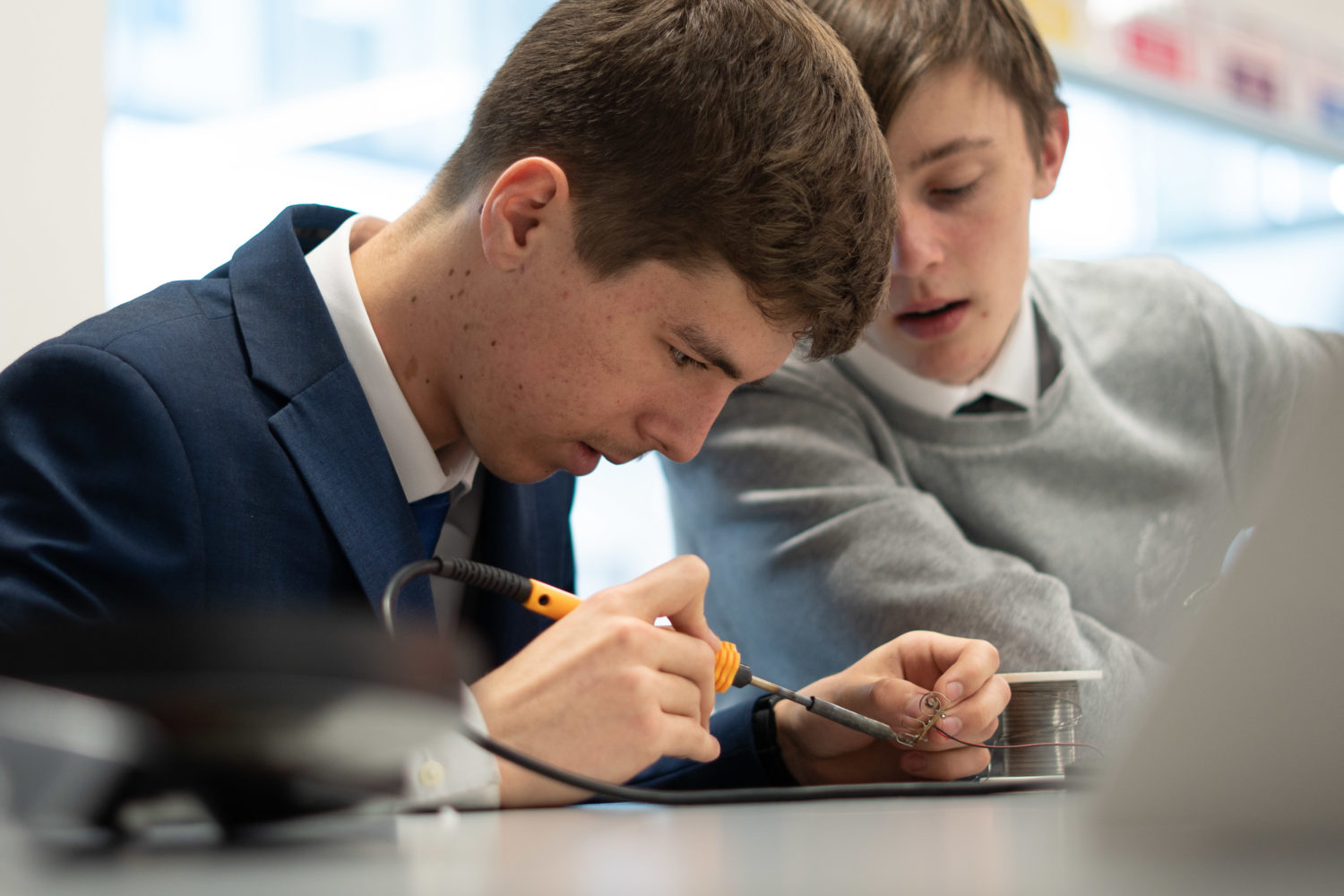 Two Leigh UTC students are shown sitting next to each other at a desk. One is shown using a soldering iron on a piece of wire.