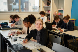 A small class of Post-16 students can be seen sat at their desks and concentrating on their work. In the foreground, a female student can be seen looking directly up at the camera.