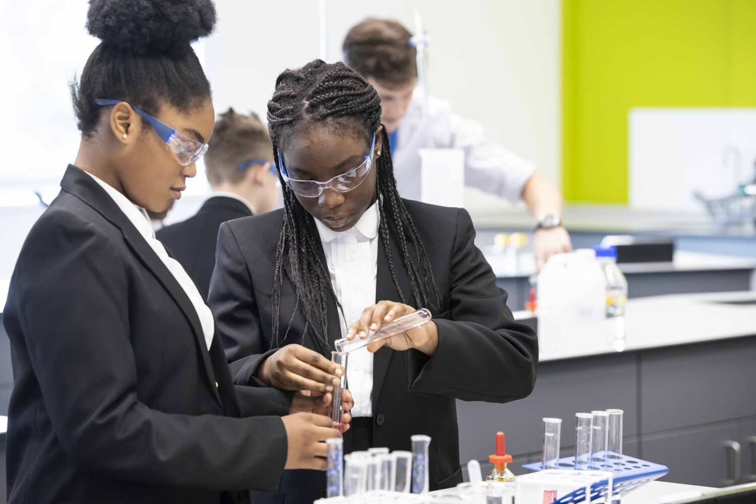 Two female students are pictured conducting a Science experiment in the foreground, using chemicals and glass vials. Other students are seen doing the same in the background.
