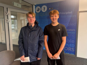 Two young men are standing side-by-side, each holding white papers or envelopes. They are both smiling, indicating positive outcomes. The background includes a blue wall with words like "KNOWLEDGEABLE," "Caring," "Principled," and "Risk-takers," consistent with a school environment. An "IB" logo is also visible, suggesting an International Baccalaureate program.